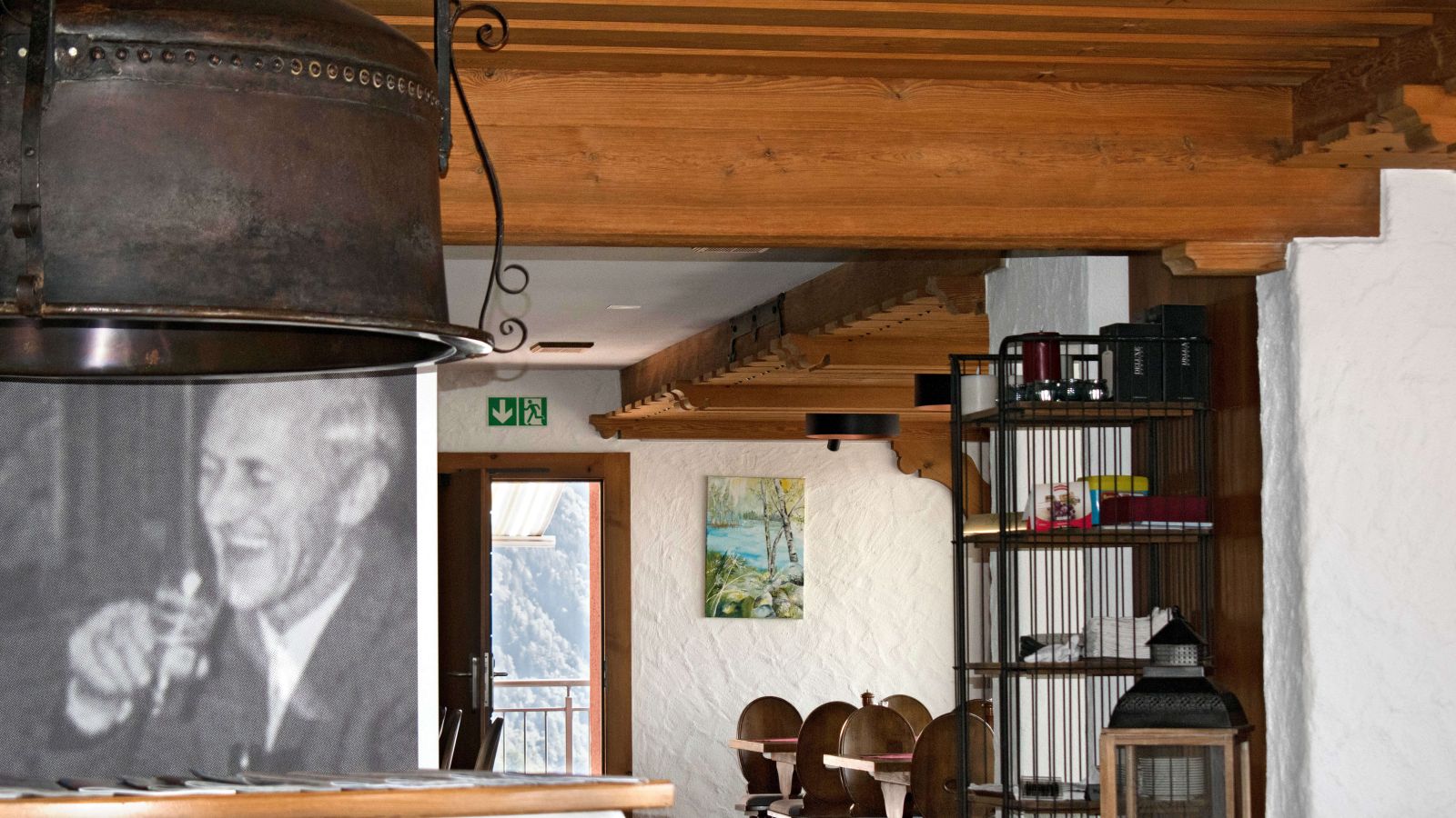 A rustic dining area featuring wooden ceiling beams, a copper lampshade, and a view of a sunny balcony.