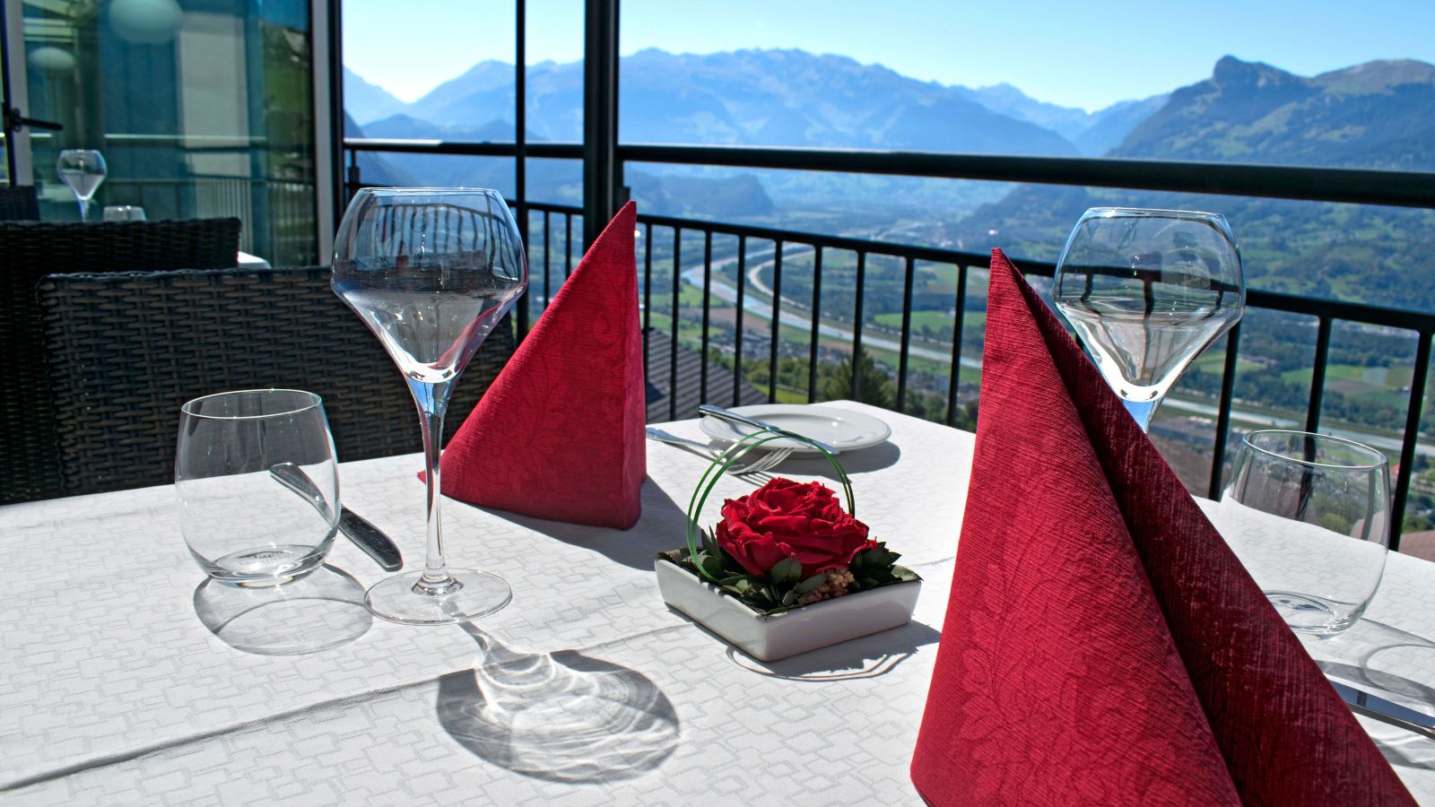 A finely set restaurant table with red napkins and a rose overlooking a stunning mountain panorama.